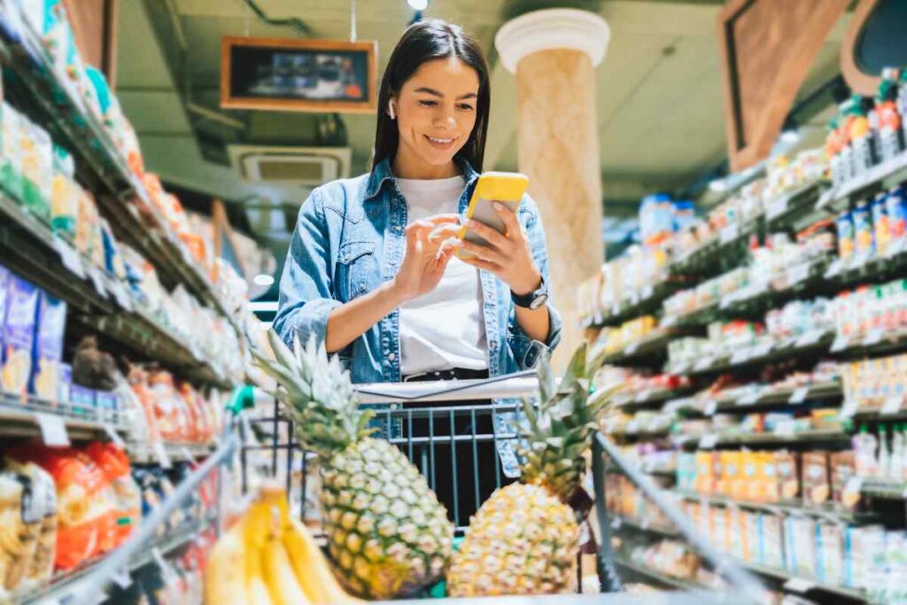 Ragazza con carrello pieno di frutta che consulta l'app yoka dallo smartphone per fare la spesa