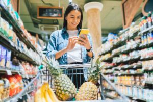 Ragazza con carrello pieno di frutta che consulta l'app yoka dallo smartphone per fare la spesa